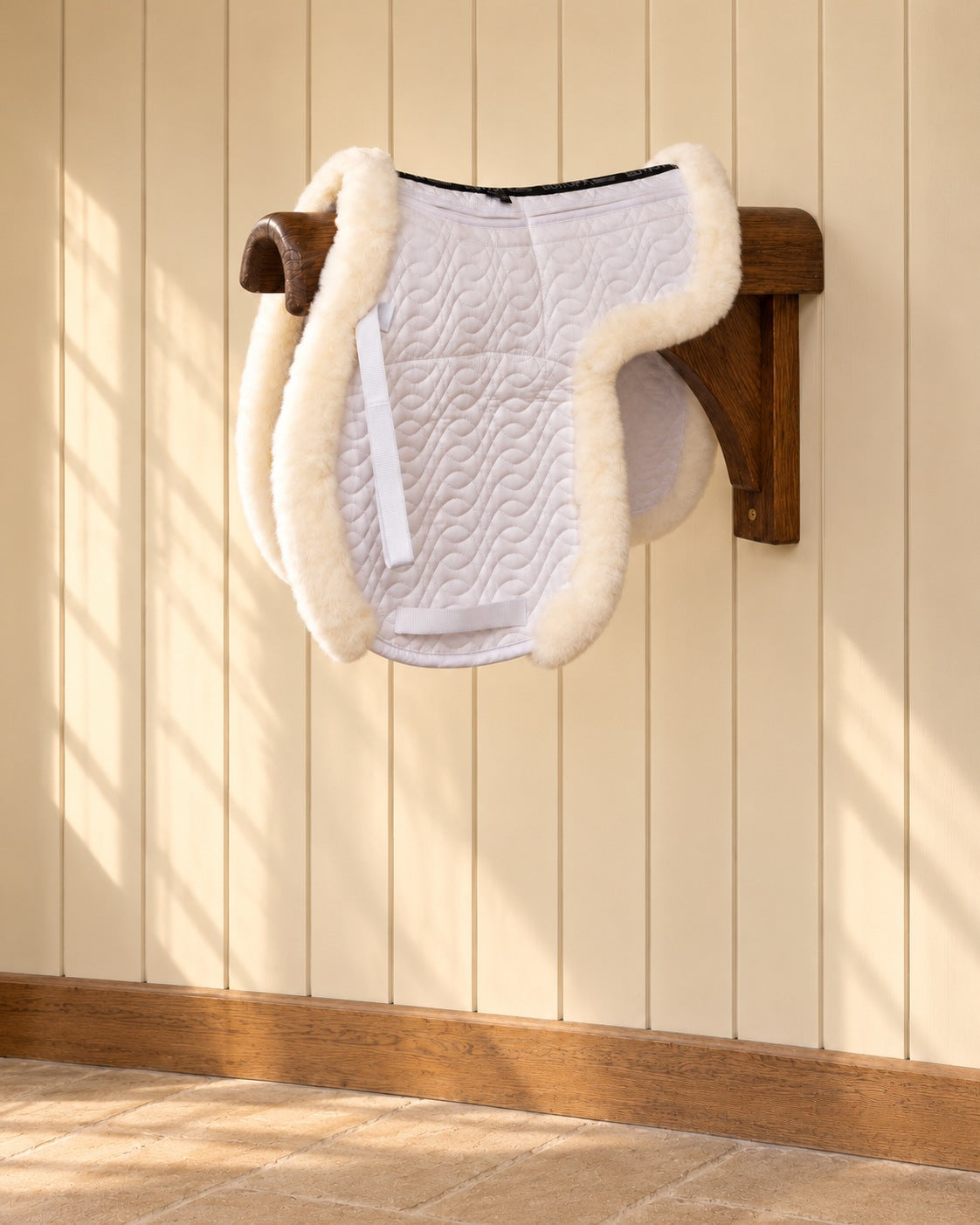 White 4-shim hunter show saddle pad with sheepskin trim on a wooden wall bracket against a cream panelled wall