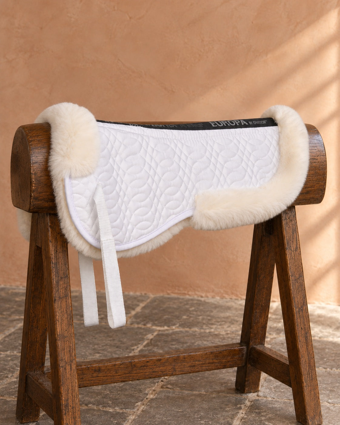 Ovation Europa sheepskin half pad in natural draped over a wooden saddle stand against a terracotta plaster wall