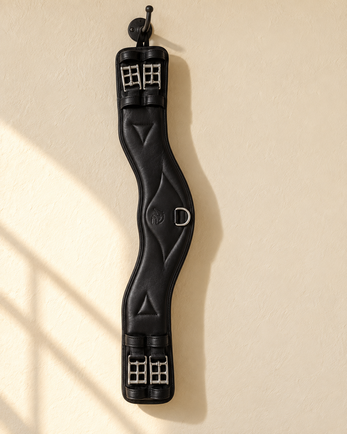 Black leather dressage girth hanging from an iron hook on a cream plaster wall, showing its anatomical curve, the centre D-ring and the two roller buckles at each end