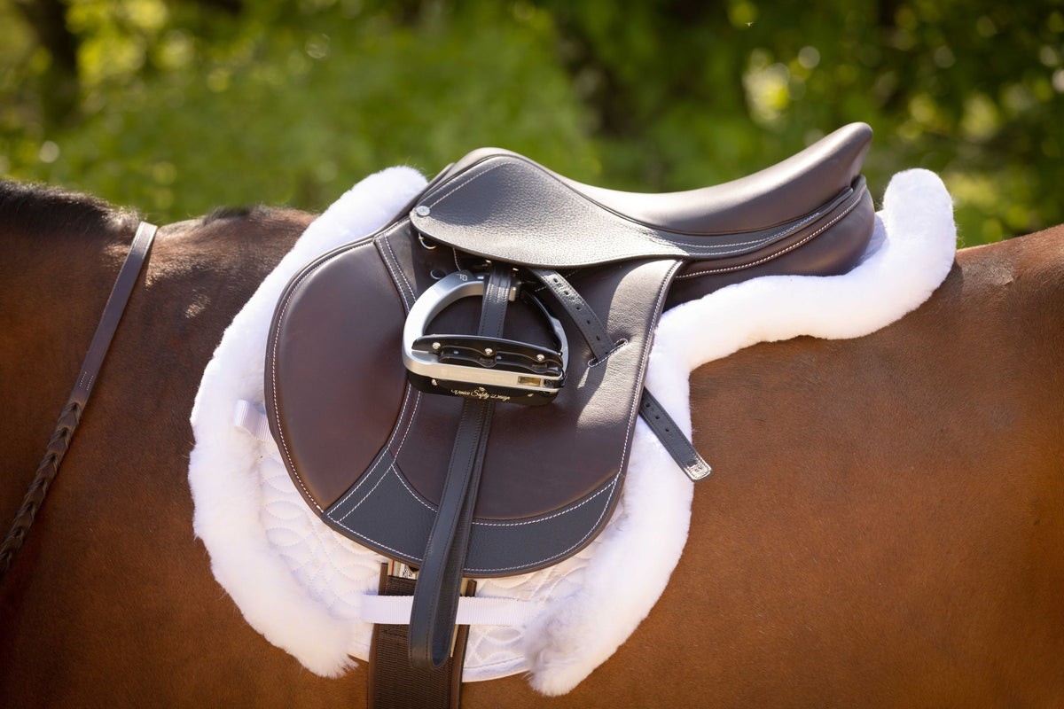 Dark brown saddle with a stirrup on a bay horse over a white fleece pad, side view outdoors