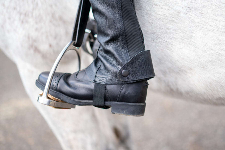 Close view of a black half chap and paddock boot in a stirrup iron, showing the ankle snap and strap under the boot