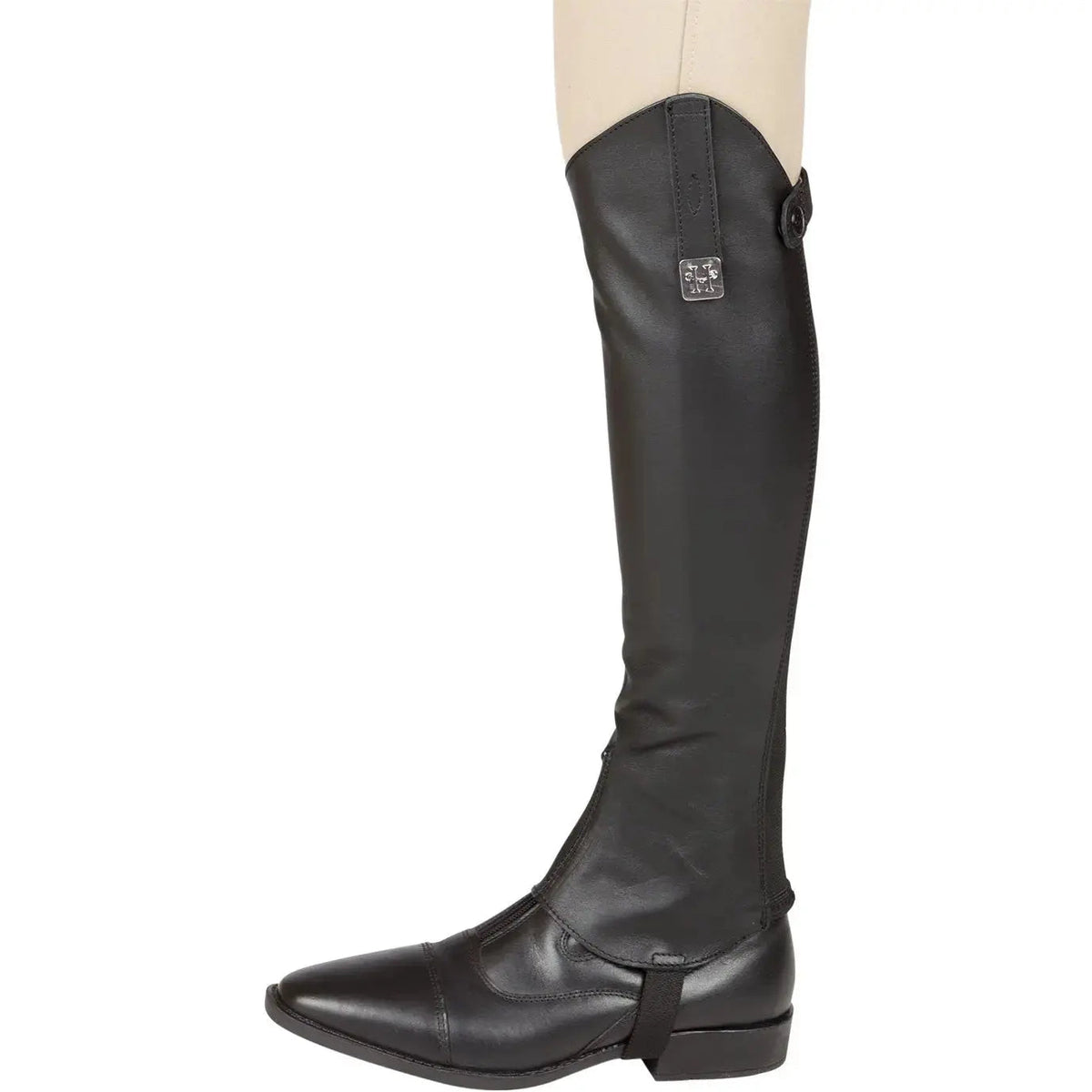 Side view of a single black leather half chap with metal logo plate and elastic strap under a black paddock boot
