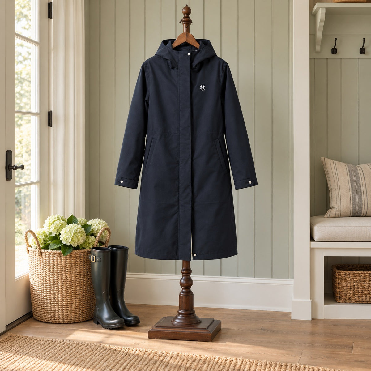 Long navy hooded rain coat with an H logo on a wooden valet stand beside tall boots in a mudroom