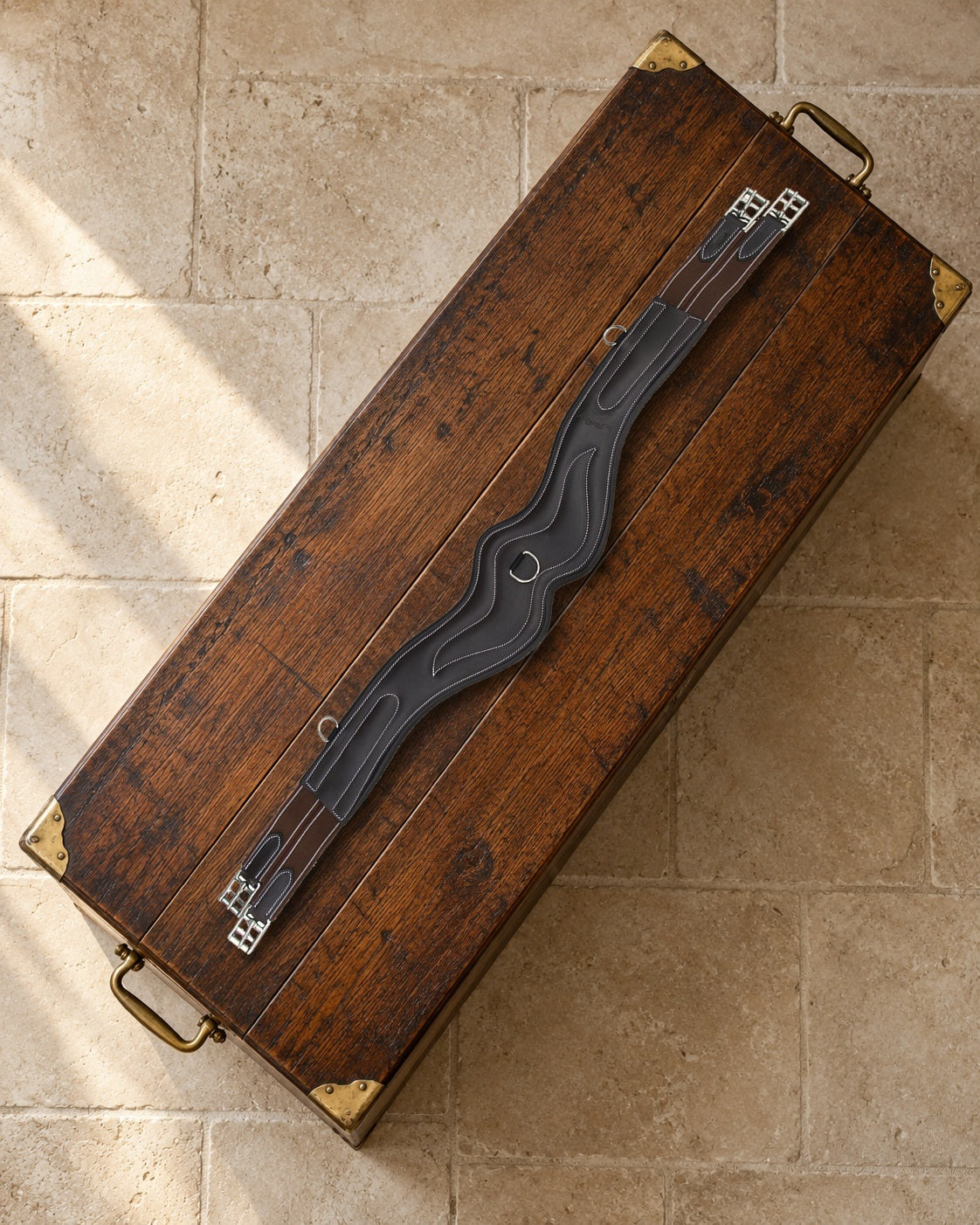 Dark brown anatomic leather girth with white stitching, brown elastic and stainless buckles lying on an oak tack trunk with brass corners