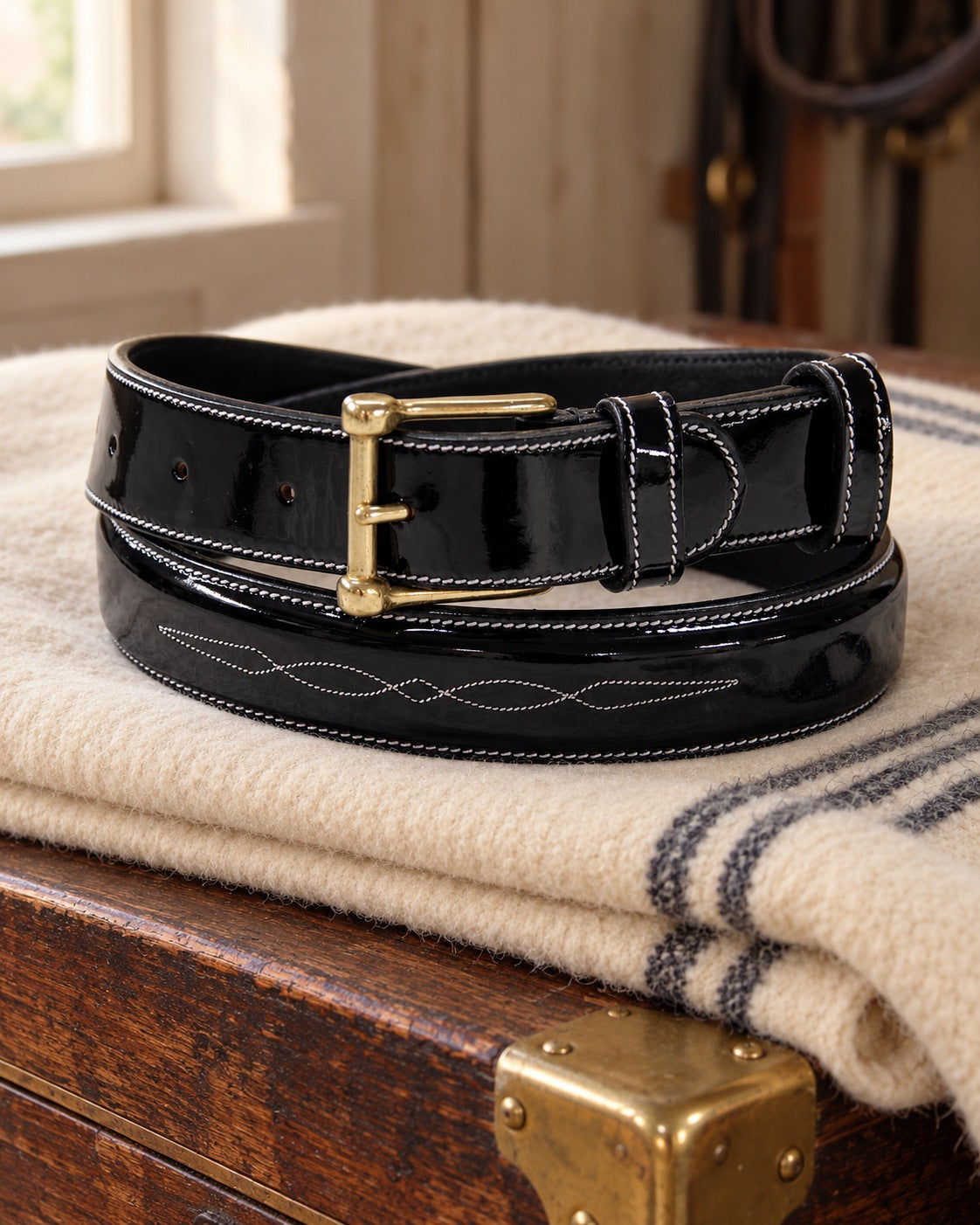 Glossy black ExionPro fancy stitched leather belt with brass buckle coiled on a wool blanket on a tack trunk