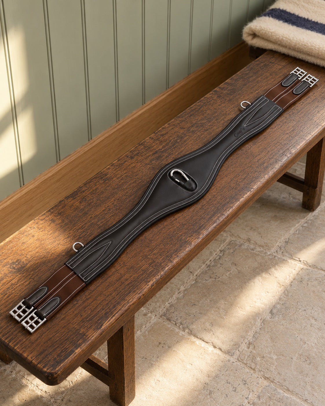 Dark leather contoured snap girth with white stitching, brown elastic ends and stainless buckles laid on an oak bench in a sage green tack room
