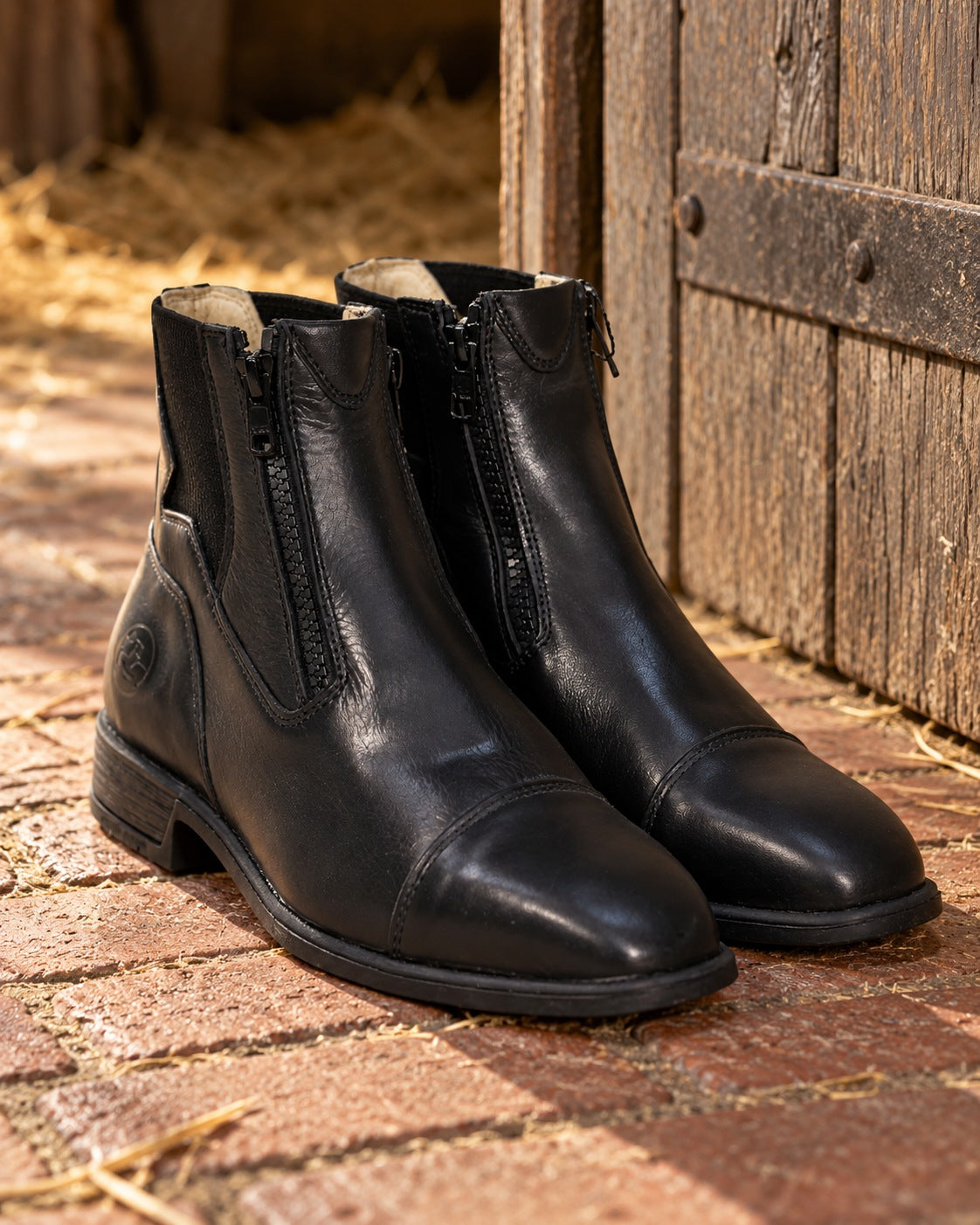 Pair of black Grand Prix Saltare Florence front zip paddock boots on a brick stable floor beside a stall door