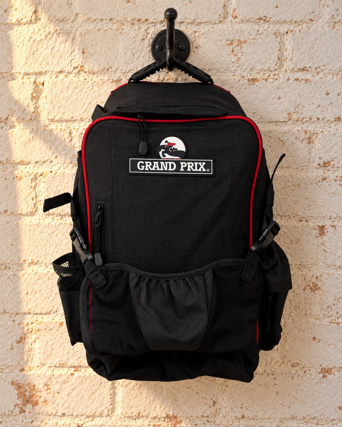 Black Grand Prix helmet backpack with red piping hanging by its rubber grab handle from an iron hook on a whitewashed brick wall