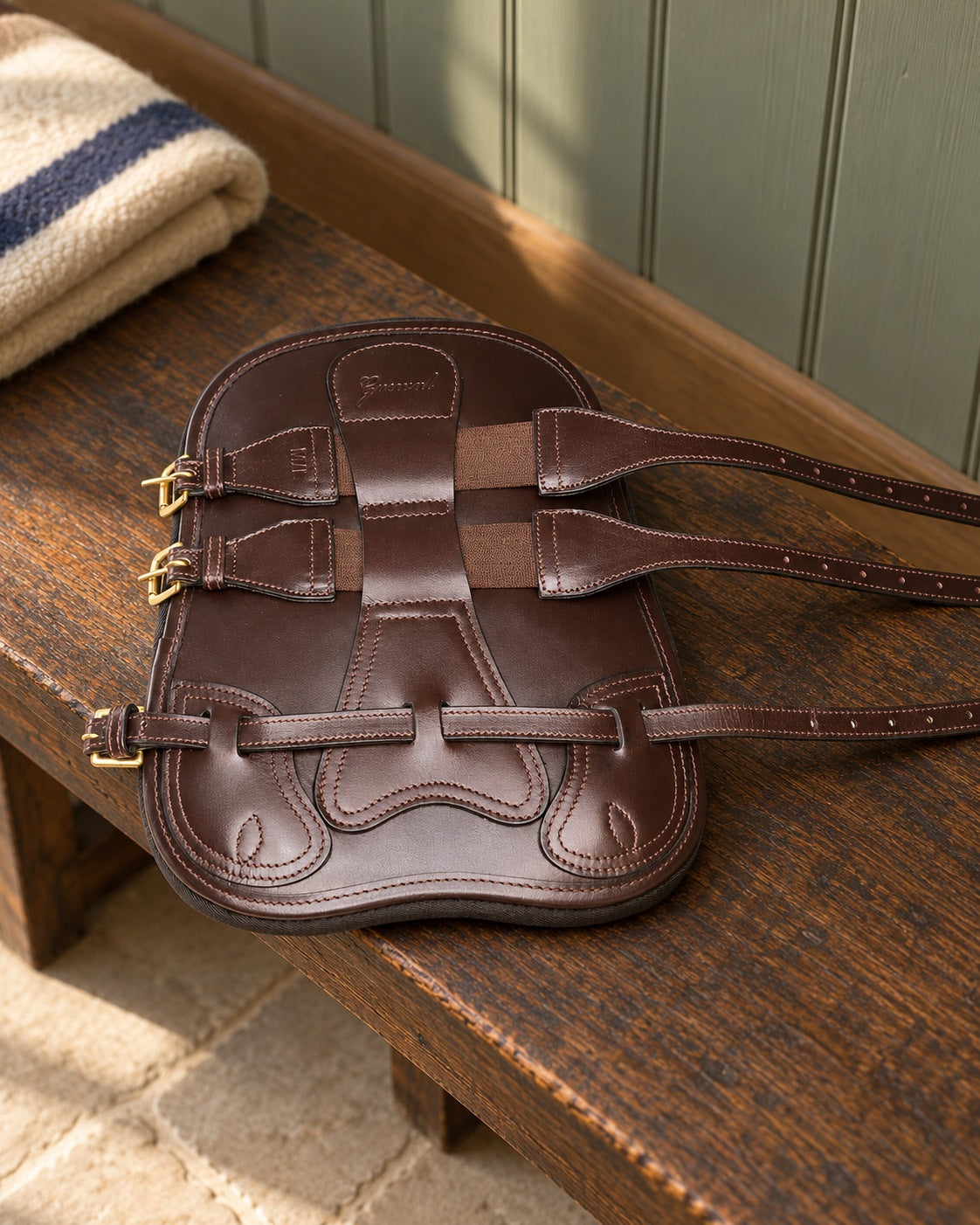 Dark brown leather open front tendon boot with brass buckles on an oak bench in a sage green tack room