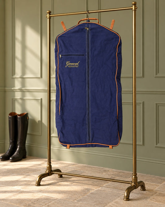 Navy canvas garment bag with tan leather piping hanging from a brass garment rail beside tall black riding boots