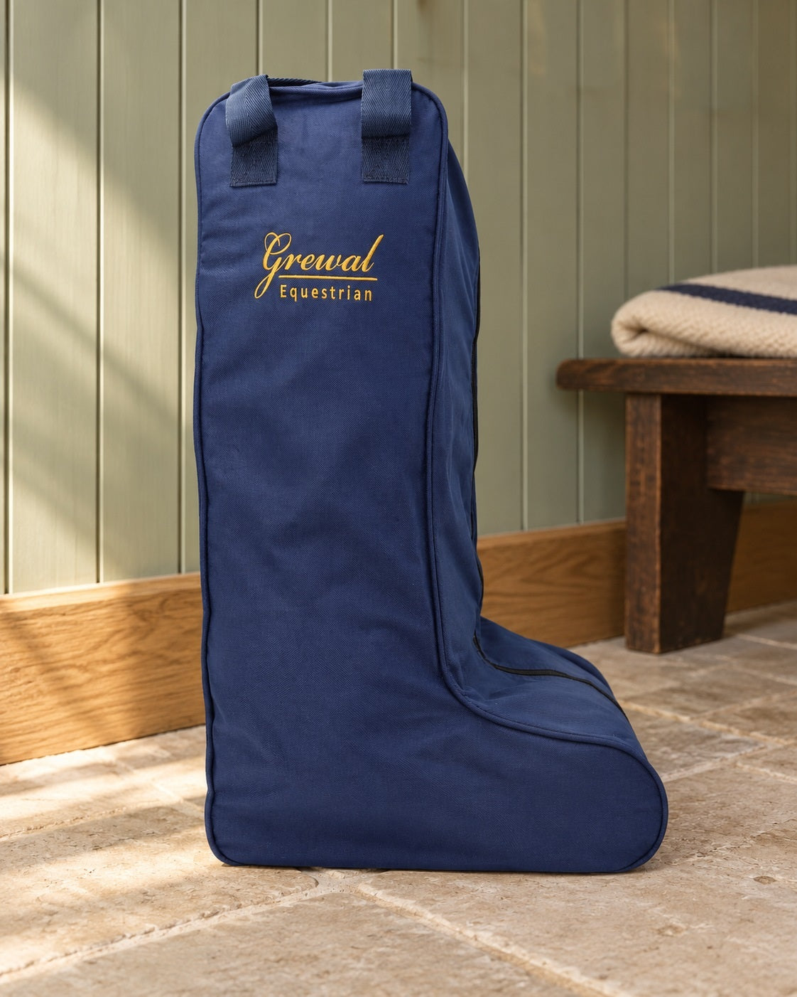 Navy canvas tall boot bag with gold embroidered logo standing on a limestone floor in a sage green tack room