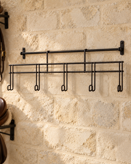 Dark wire HDR 5 hook bridle and tack rack with two rails, hanging by its two top hooks from a black iron rail on a limestone tack room wall, saddles on wall racks at the left edge