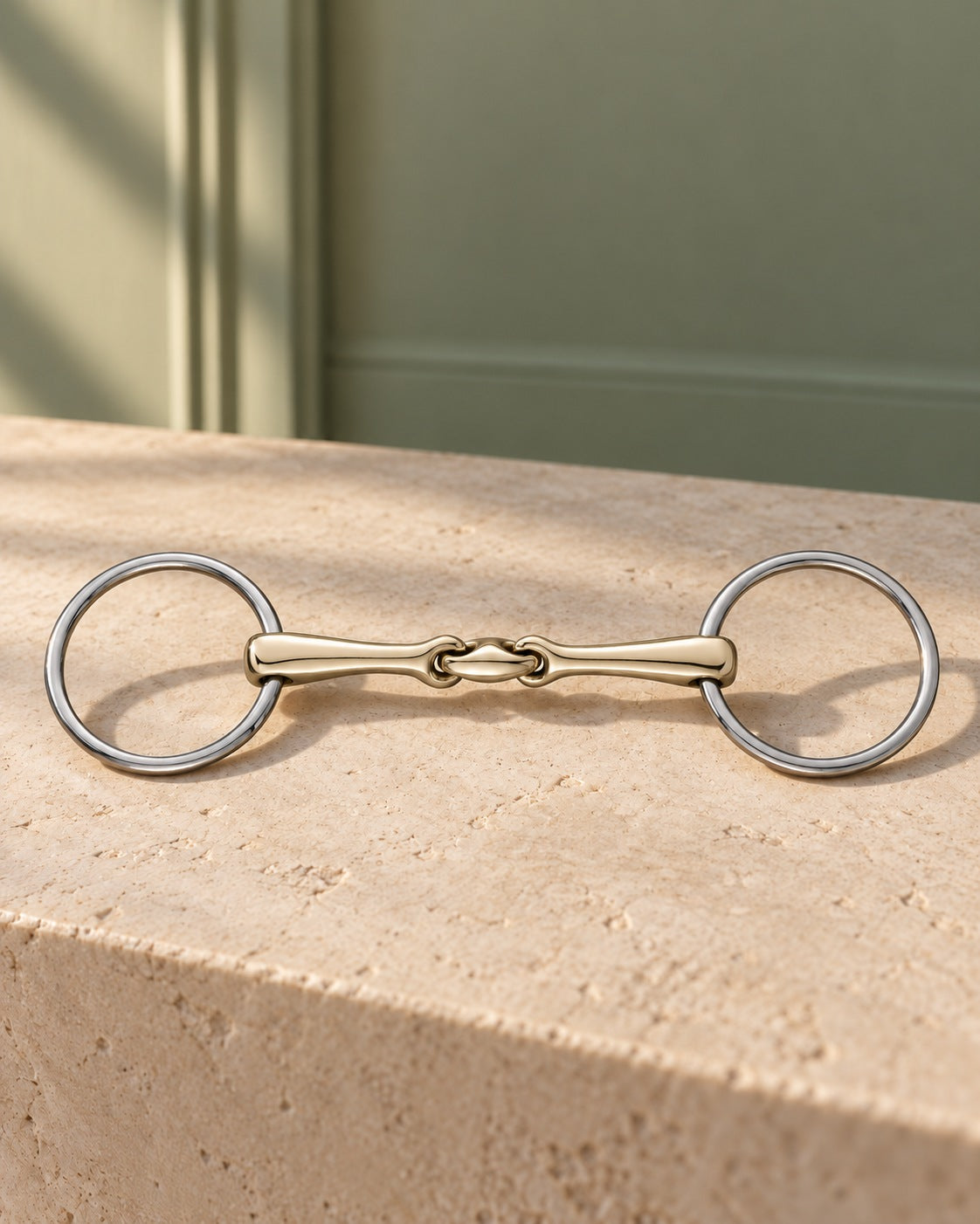 Loose ring snaffle bit with stainless steel rings and a double-jointed champagne gold mouthpiece, resting on a limestone ledge in front of a sage green wall