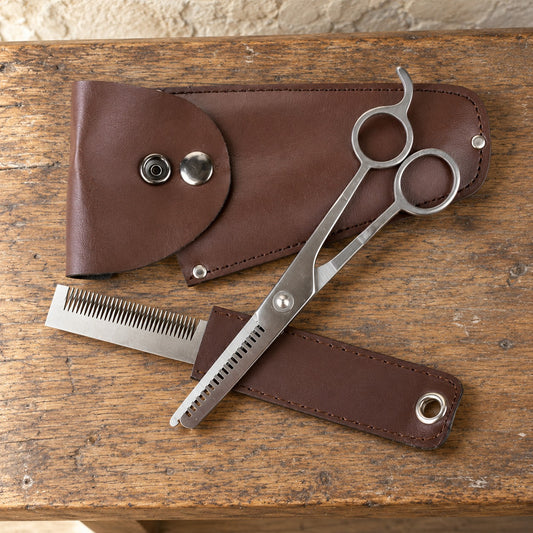 Mane and tail trim kit with thinning scissors, a thinning comb, a brown leather snap case and belt loop strip, laid flat on an oak tack room bench