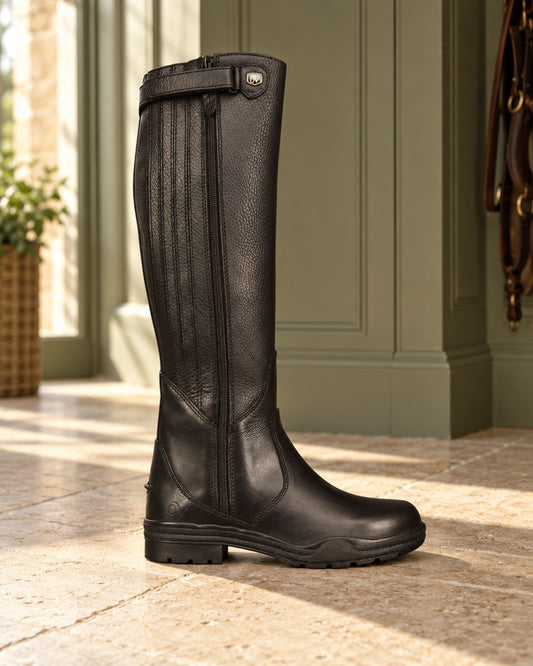 Black leather waterproof tall riding boot with a pebbled grain shaft, vertical elastic gusset panels, a full-length zip and a lugged rubber sole, standing on a limestone floor beside sage green panelling