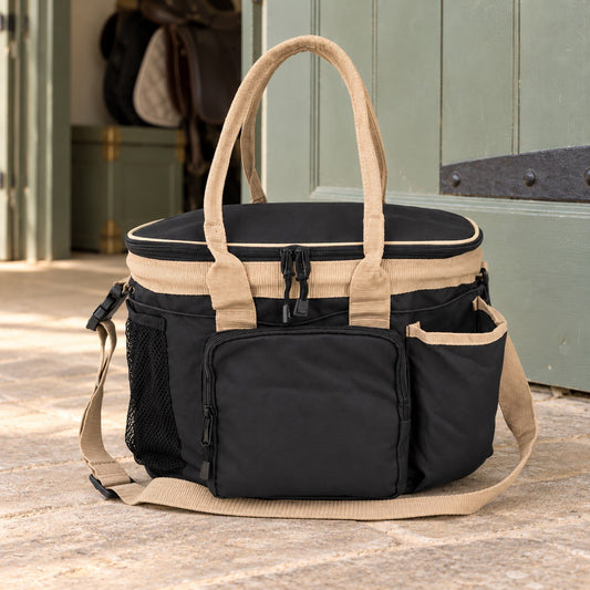 Multi-pocket grooming tote in black with beige handles, lid trim and shoulder strap, front zip pocket and mesh side pocket, on a limestone stable aisle floor beside a sage stable door