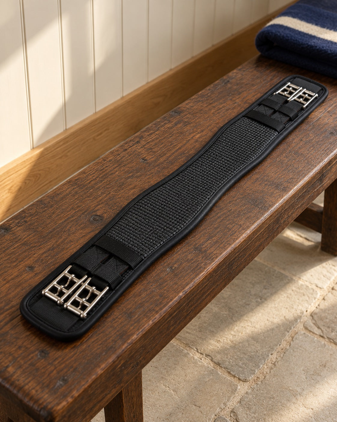 Black textured dressage girth with stainless double buckles at both ends laid flat on an oak bench in a cream tack room