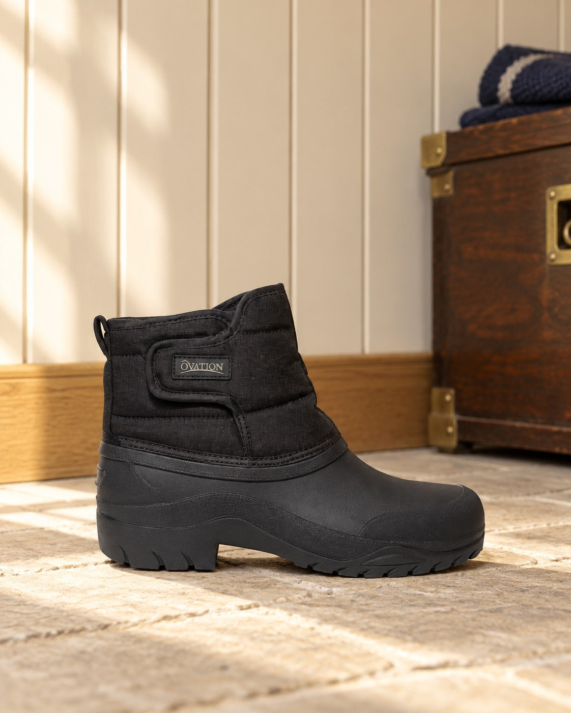 Black quilted winter paddock boot with a hook-and-loop front strap standing on a limestone floor beside an oak tack trunk