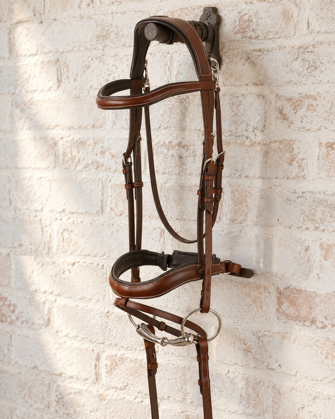 Brown Ovation Cambridge anatomic bridle with loose ring bit and reins hanging from a cast iron bridle bracket on a whitewashed brick wall