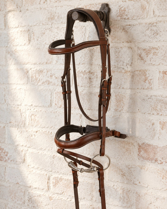 Brown Ovation Cambridge anatomic bridle with loose ring bit and reins hanging from a cast iron bridle bracket on a whitewashed brick wall