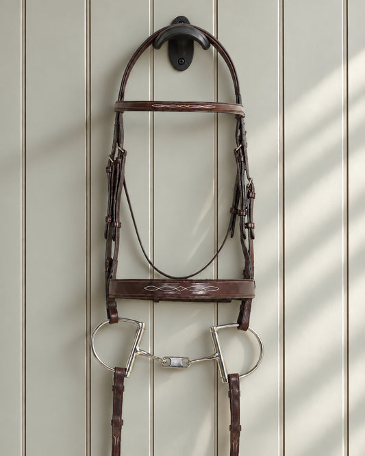 Ovation Classic wide noseband bridle with reins hung on a black cast iron bridle bracket against a grey panelled wall