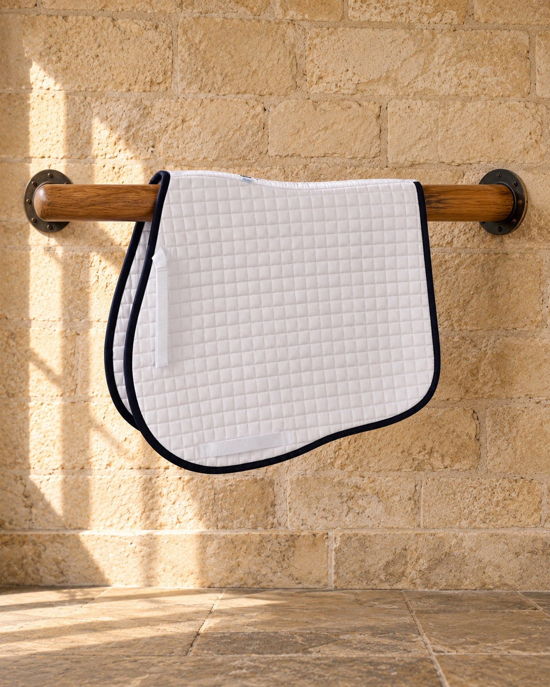 White Coolmax jumper saddle pad with navy piping on a wooden rail against a limestone wall