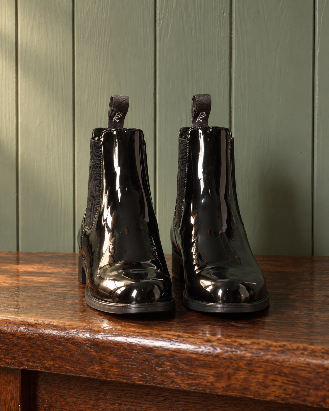 Pair of black patent leather Ovation Finalist jodhpur boots on an oak bench against a sage green plank wall