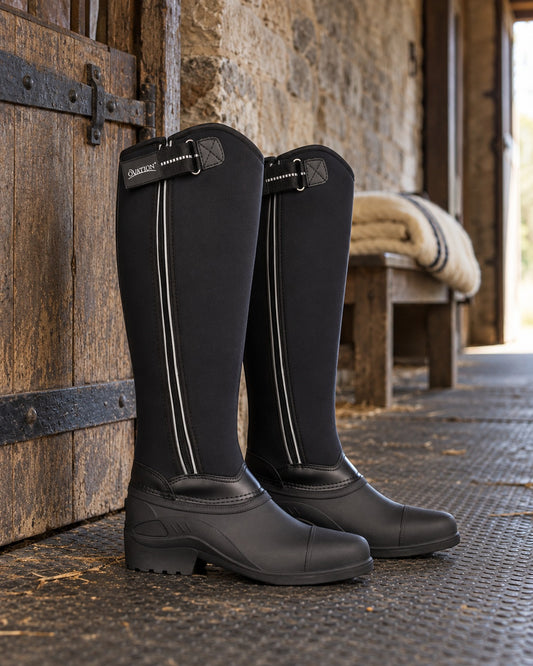 Pair of black Ovation Highlander tall winter riding boots with reflective zip piping in a stone barn aisle