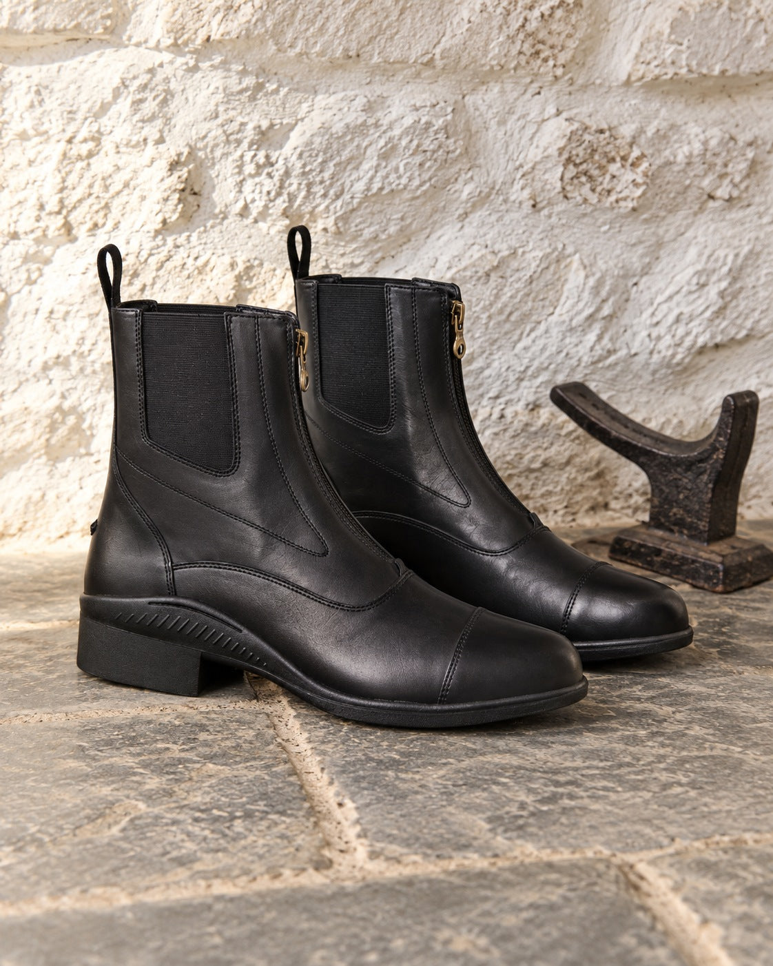 Pair of black Ovation men's Aeros Showmaster zip paddock boots on flagstone beside a cast iron boot jack