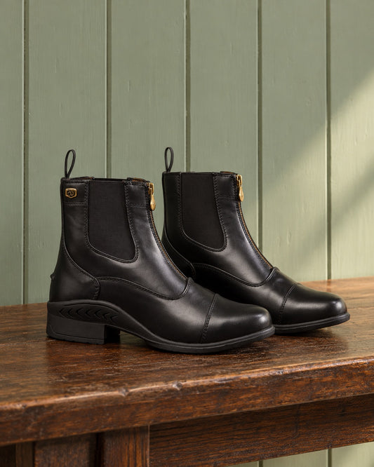 Pair of black Ovation Quantum zip paddock boots on an oak bench against a sage green plank wall