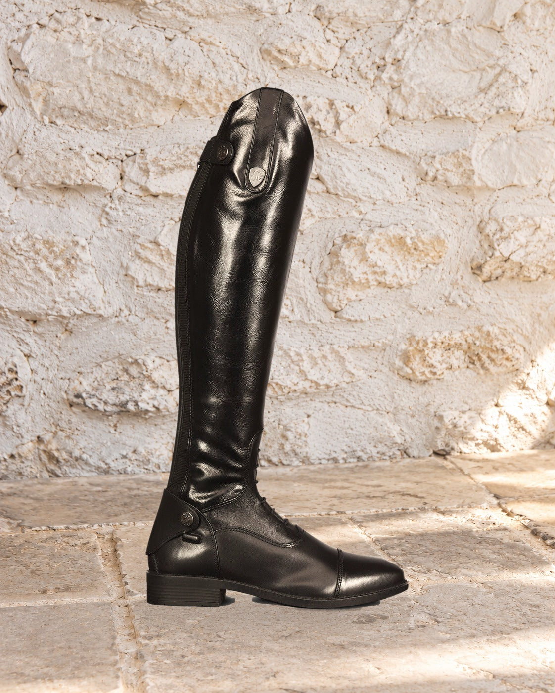 Black Ovation Sofia Grip tall riding boot in side profile on a limestone floor against a whitewashed stone wall