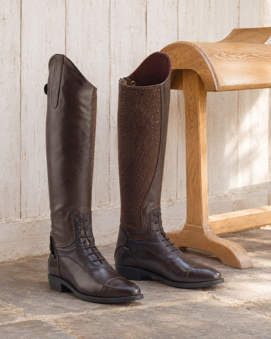 Pair of brown Ovation Sofia Grip tall boots with horseshoe pattern grip panel beside an oak saddle stand