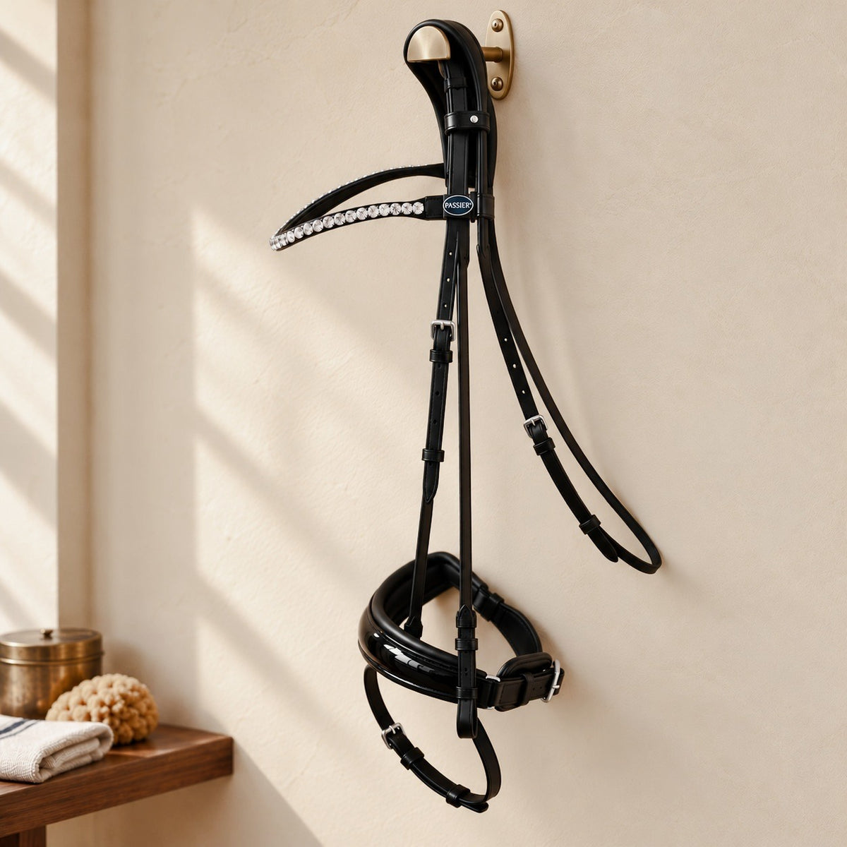 Passier Harmony snaffle bridle in black leather with a crystal browband and patent noseband, hanging from a brass bridle hook on a warm cream plaster tack room wall