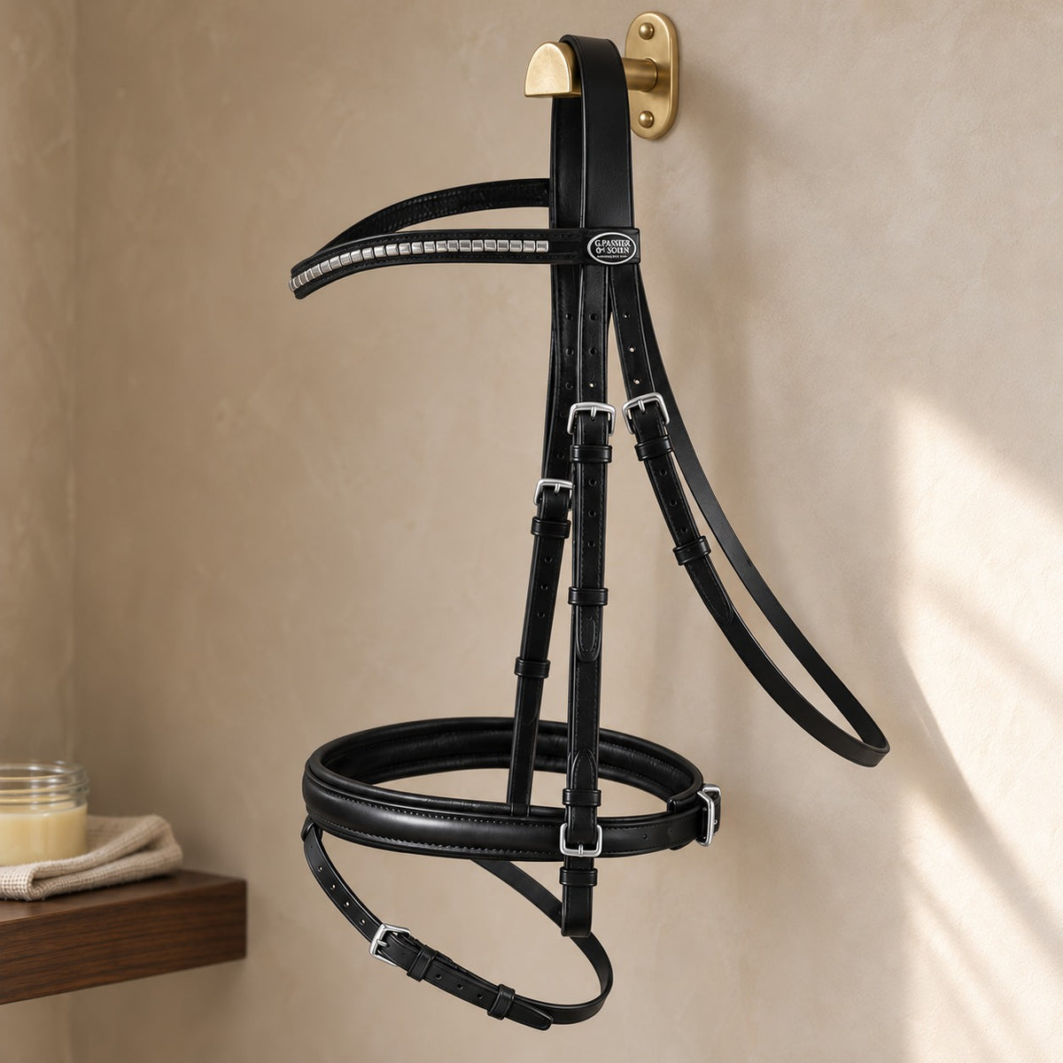 Passier Juno snaffle bridle in black leather with a silver clincher browband and padded flash noseband, hanging from a brass bridle hook on a taupe polished plaster tack room wall