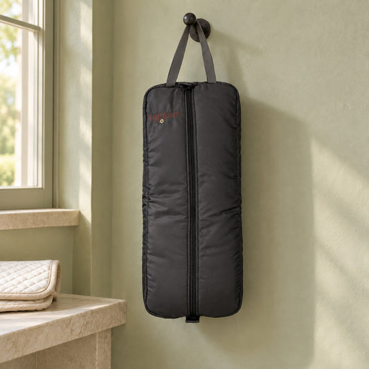 Charcoal padded Tabelo halter and bridle bag with a full-length center zip, grey carry handle and small red embroidered logo, hanging from an iron peg on a pale sage tack room wall