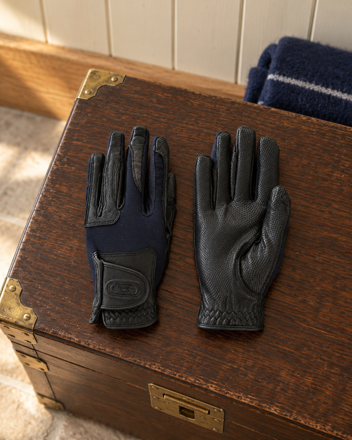 Pair of black and navy riding gloves, one showing the perforated leather palm, on an oak tack trunk with brass corners