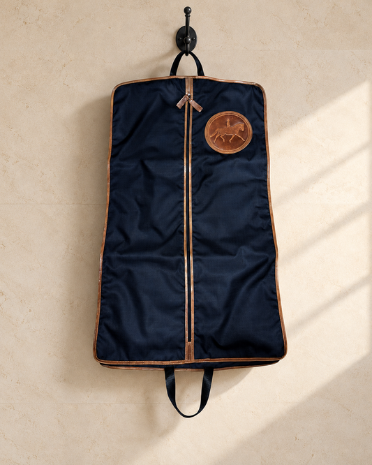 Navy Tryon Dressage long garment bag with tan leather binding, centre zip and an embossed dressage horse and rider medallion, hanging from a black iron hook on a pale limestone tack room wall