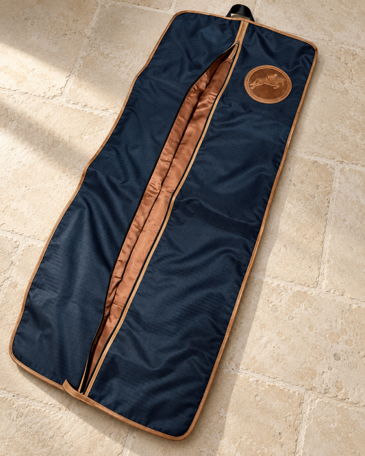 Navy Tryon Hunter/Jumper long garment bag with tan leather binding, zip partly open to the tan lining and an embossed jumping horse medallion, laid flat on a pale limestone tack room floor
