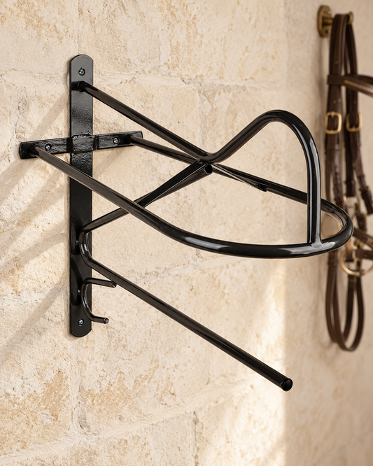 Black metal wall saddle rack with a curved saddle bar, two small tack hooks and a long lower bar, mounted on a limestone tack room wall beside a hanging bridle