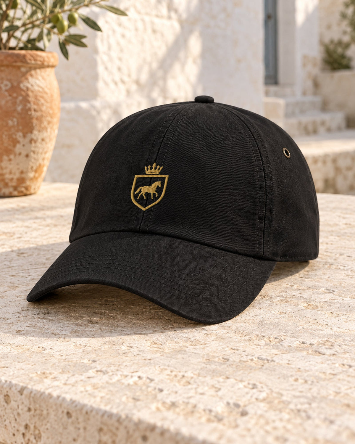 Canter & Crest Signature Crest Cotton Cap in Black, gold embroidered Signature crest on the front panel, on a pale limestone table in a whitewashed courtyard