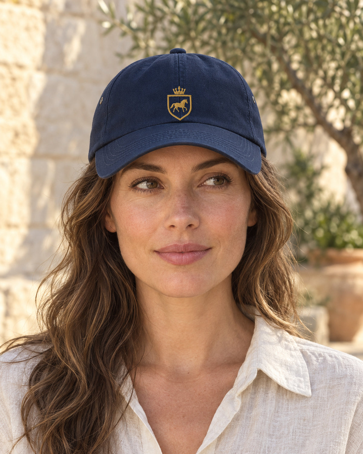 AI-generated image. Woman wearing the Canter & Crest Signature Crest Cotton Cap in Navy in a Mediterranean courtyard, gold embroidered Signature crest on the front panel