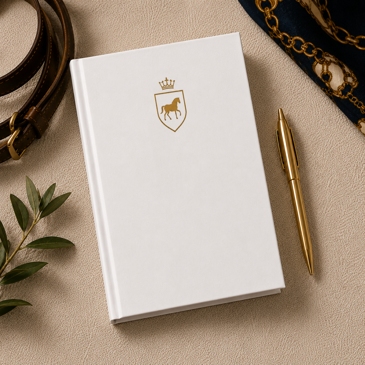 White hardcover journal with a gold crest of a horse under a crown, flat lay with a gold pen and belt