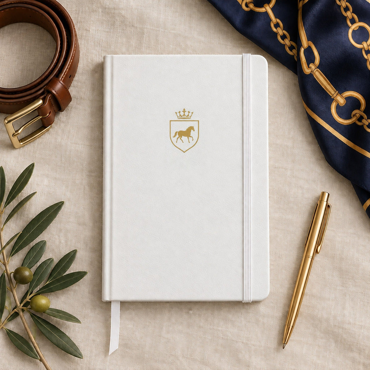 White hardcover notebook with a gold horse and crown crest, styled with a brown belt, scarf and gold pen