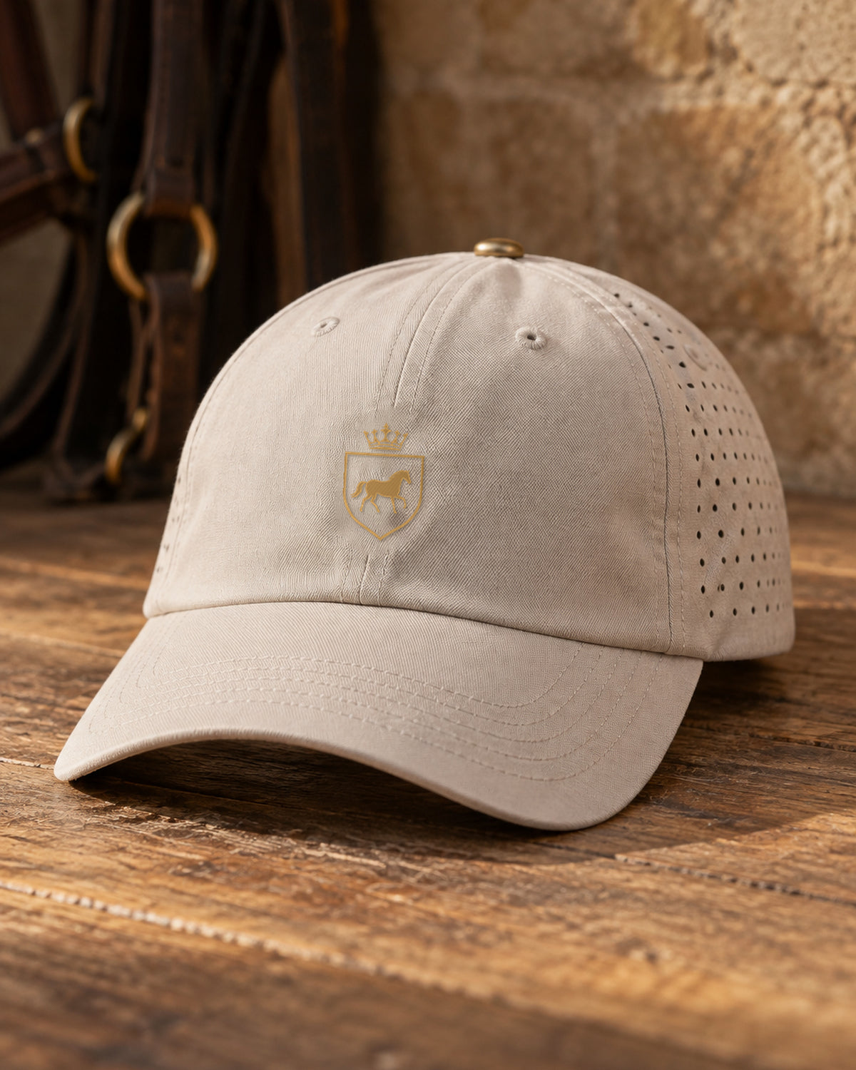 Canter & Crest Signature Crest Perforated Cap in Stone, gold embroidered Signature crest on the front panel, resting on weathered timber with leather bridles behind
