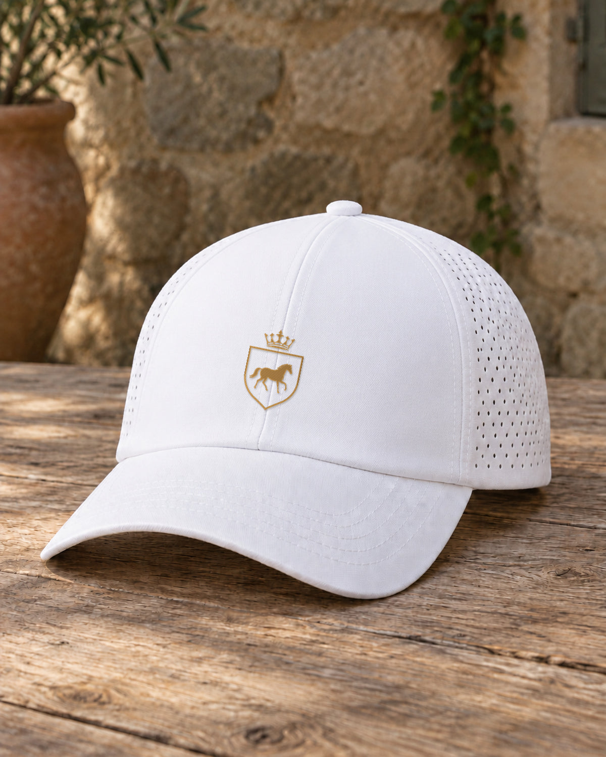 Canter & Crest Signature Crest Perforated Cap in White, gold embroidered Signature crest on the front panel, resting on a sun-bleached olive-wood table in a Provencal courtyard