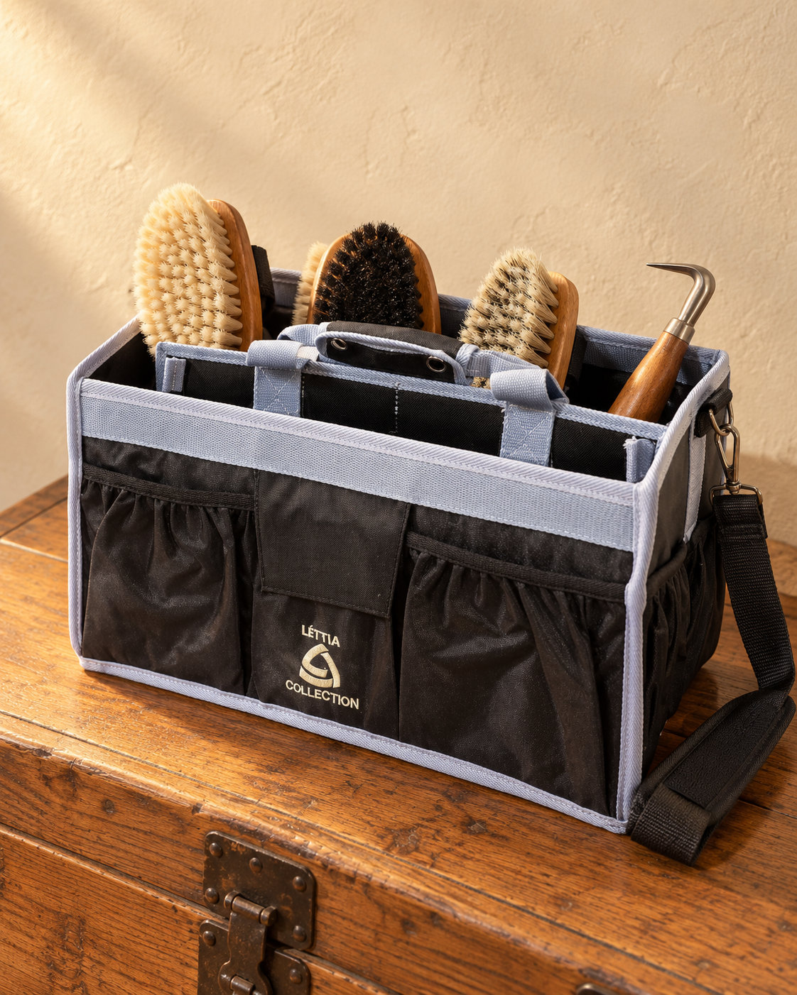 Black nylon grooming tote with light grey webbing trim on a wooden tack trunk, holding wooden-backed body brushes and a hoof pick