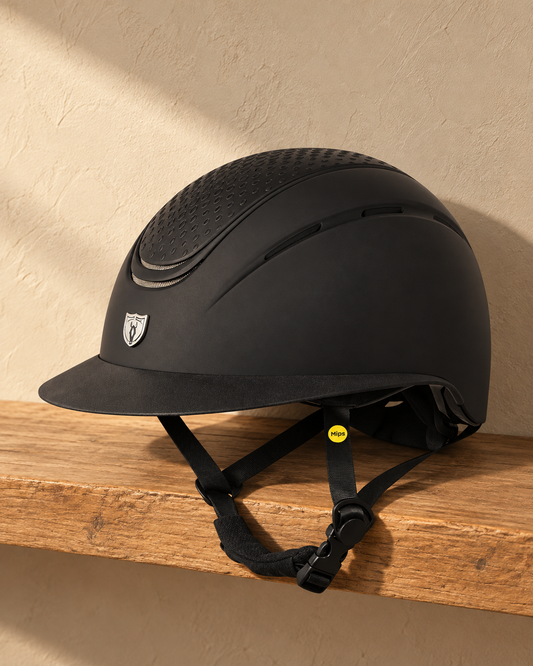 Matte black riding helmet on an oak shelf, showing the dimpled top vent panel, silver shield emblem and black harness with the yellow Mips tab