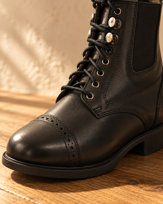 Close view of a black leather paddock boot, punched brogue cap toe, round eyelets, silver hooks and a black elastic side gore