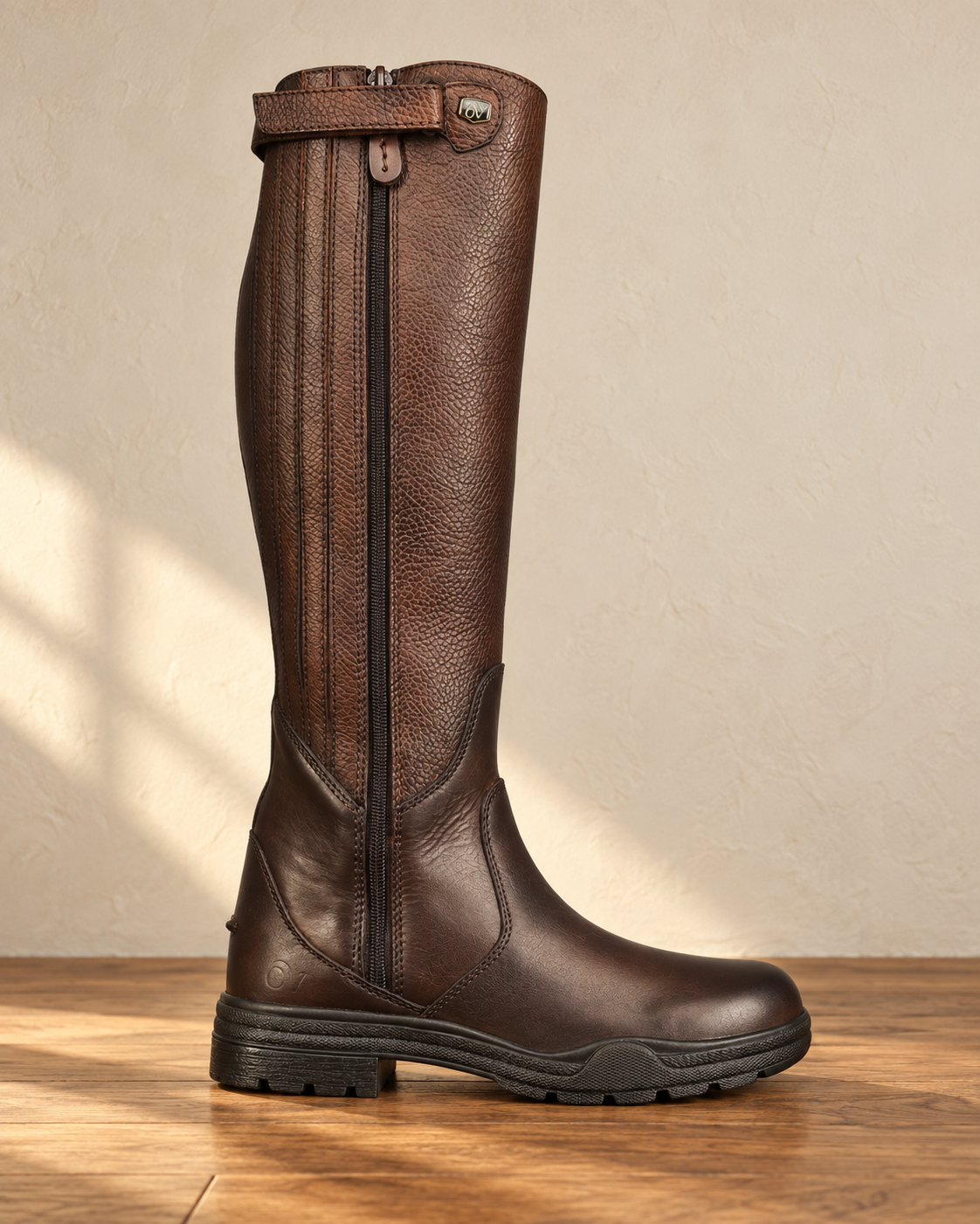 Brown tall riding boot standing on an oak floor, pebbled grain shaft, smooth leather foot, full-length zip with a guard and a lugged rubber sole