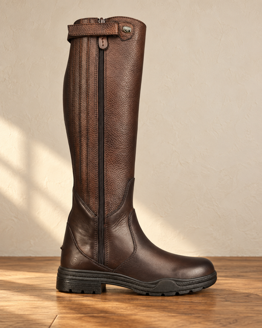 Brown tall riding boot standing on an oak floor, pebbled grain shaft, smooth leather foot, full-length zip with a guard and a lugged rubber sole